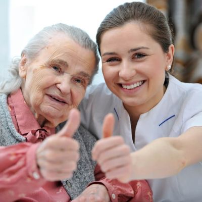 Eine Seniorin und eine weibliche Pflegekraft im Kassack strecken ihre Daumen nach oben.