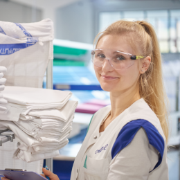 Die Qualitätsmanagerin kontrolliert einen Stapel Mietwäsche auf Hygiene mit Laborkittel und Schutzbrille.