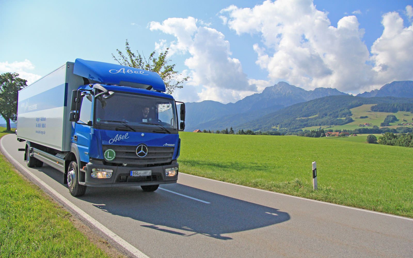 Blautransparent überlagert - Abel-LKW mit blauem Führerhaus und weißem Kofferaufbau fahrend auf der Teerstraße vor grünen Wiesen und Bergpanorama im Berchtesgadener Land.