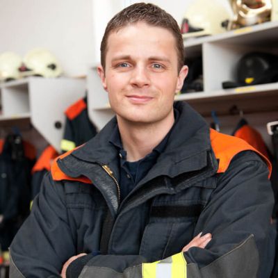 Ein Feuerwehrmann lächelt mit verschränkten Armen in Schutzausrüstung mit Leuchtstreifen in die Kamera.